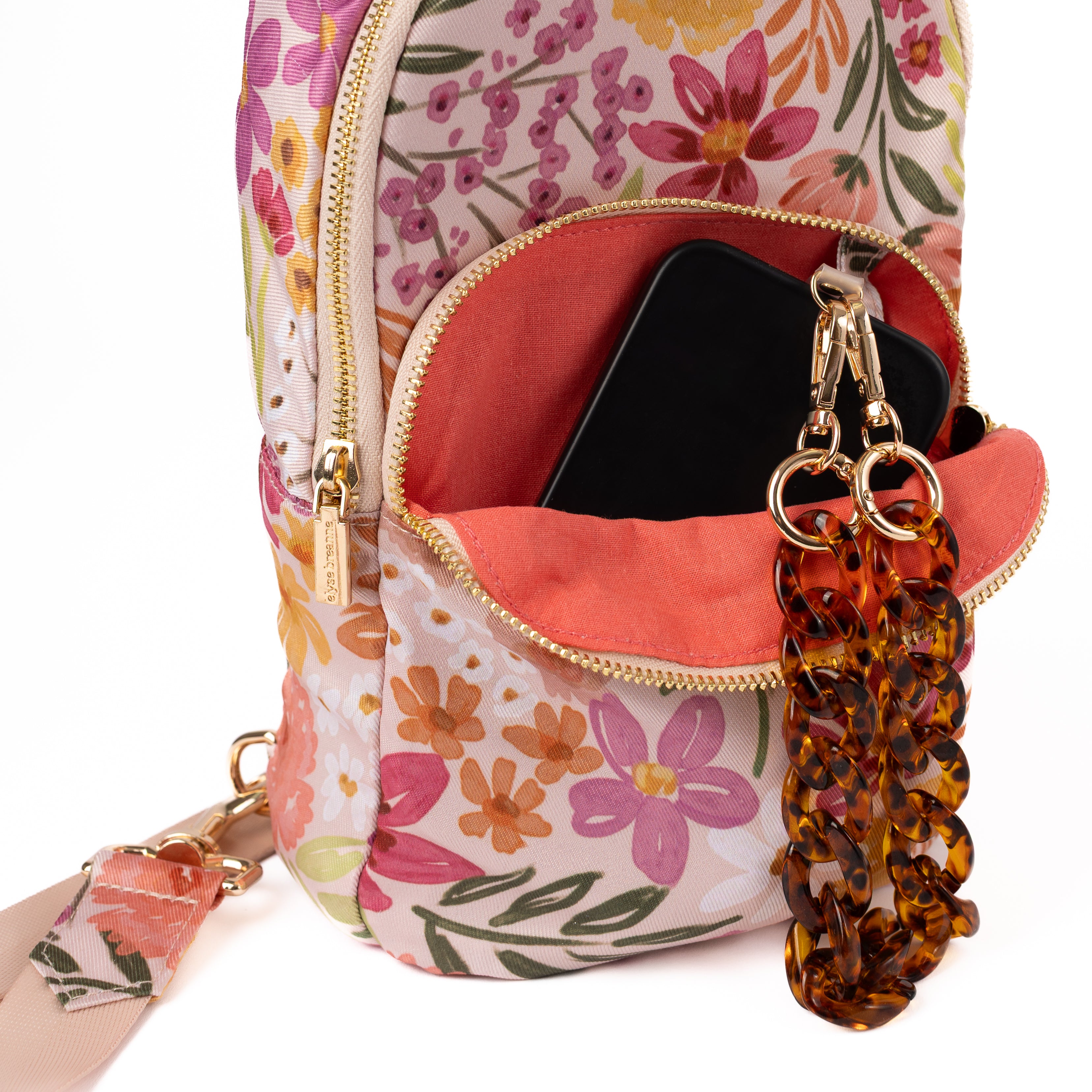 A close-up of a floral patterned backpack or shoulder bag with gold-tone hardware, shown open to reveal a coral-pink interior and a black phone inside. A tortoiseshell chain keychain is attached to the zipper.