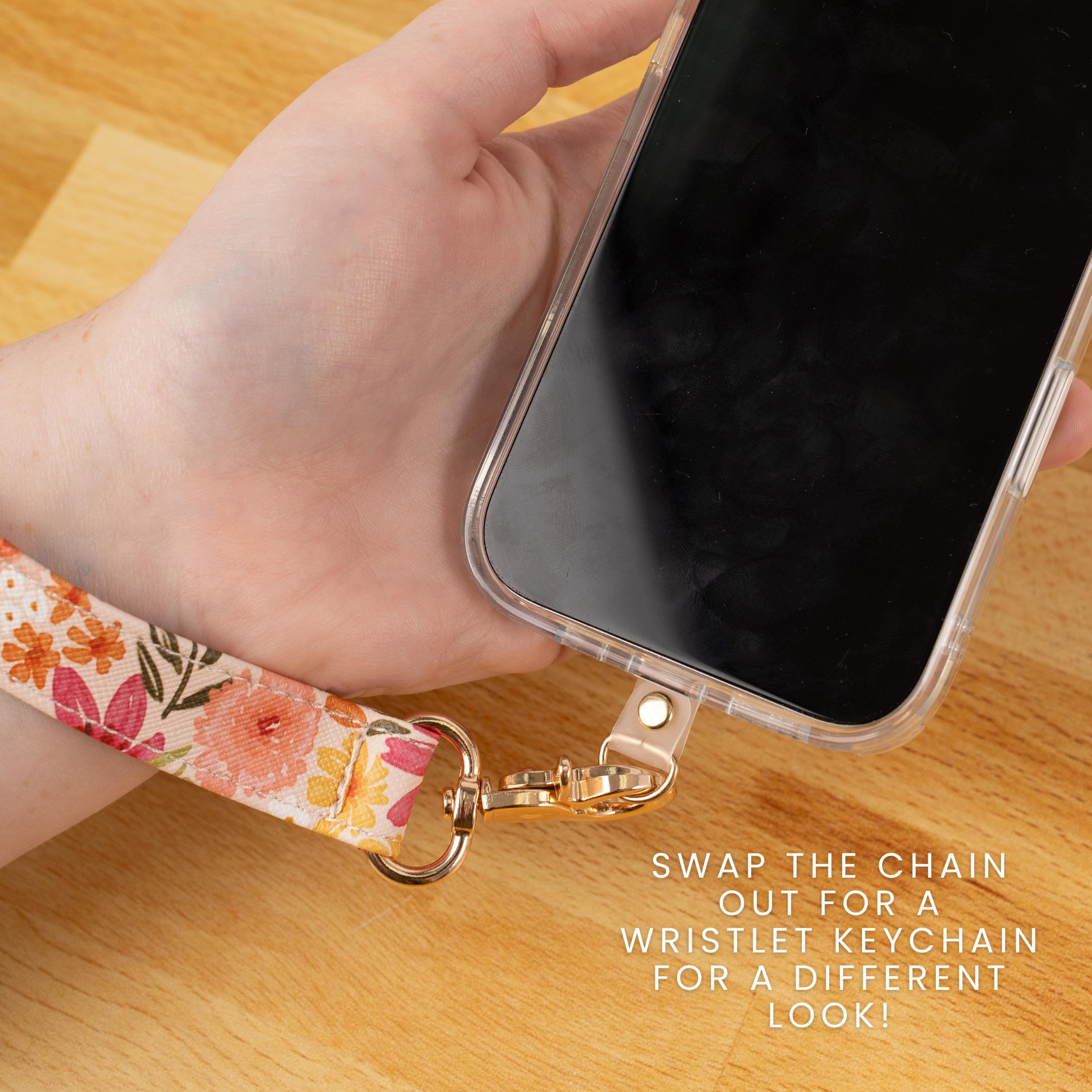 A hand holds a clear smartphone case with a floral wristlet keychain attached via a gold-tone clasp, shown on a light wood surface. White text reads, 'SWAP THE CHAIN OUT FOR A WRISTLET KEYCHAIN FOR A DIFFERENT LOOK!'.